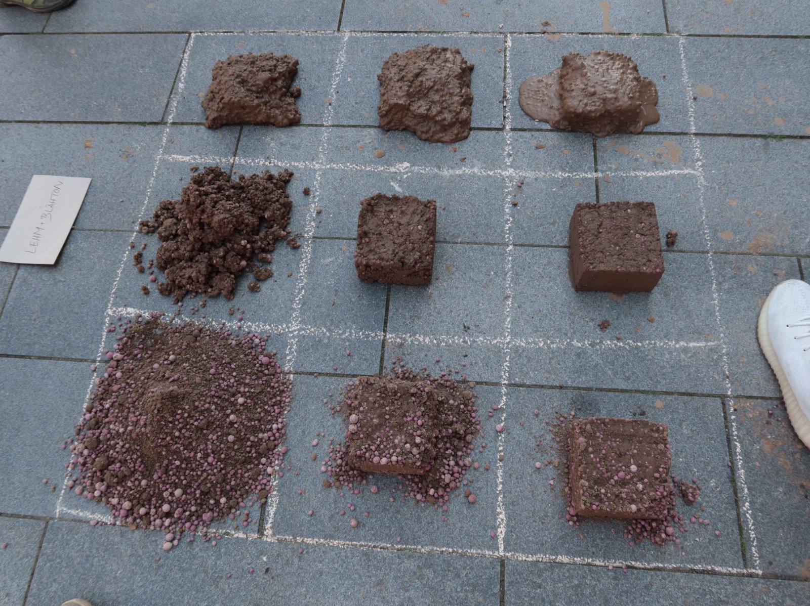 Lehmprobenraster: neun Lehmwürfel mit verschiedenen Mischungen auf Pflasterstein