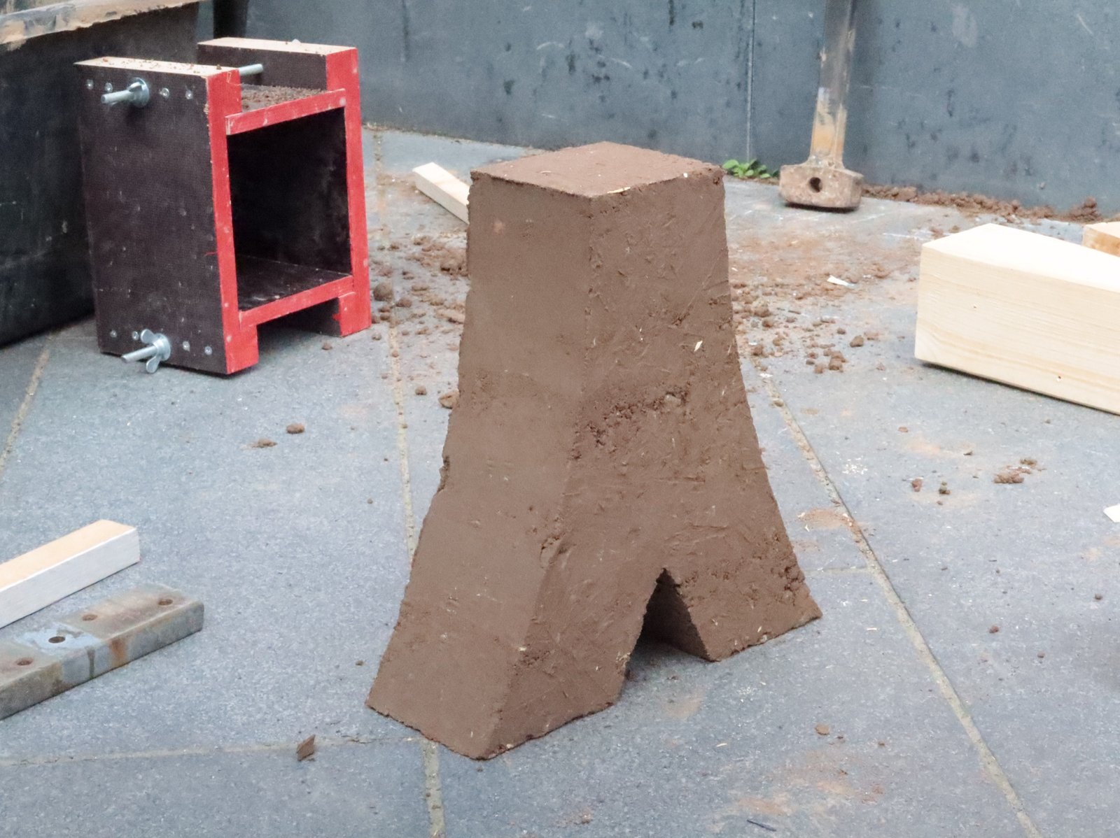 Stampflehm A-Form: frisch entformter Lehmkörper neben roter Schalung auf Pflaster